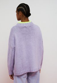 Donna con capelli intrecciati che indossa un maglione lavorato a maglia color lavanda con dettaglio del colletto verde neon e pantaloni viola chiaro, vista di spalle.