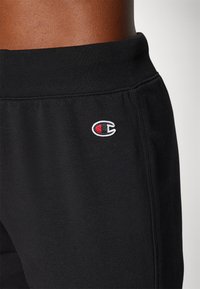 Schwarze Jogginghose aus weichem Stoff mit einem gerippten Taillenbund. Verfügt über ein kleines, gesticktes Champion-Logo in Rot und Weiß.