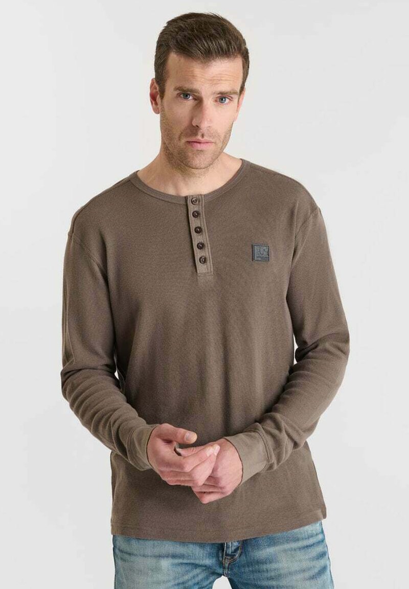 Hombre con camiseta de manga larga color marrón tipo henley con botón y vaqueros azules, de pie contra un fondo claro y liso.