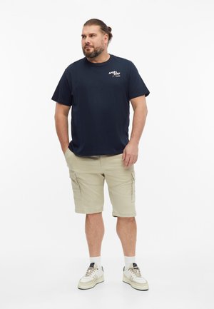 Mężczyzna z brodą i związanymi do tyłu włosami stoi w granatowym t-shircie, beżowych spodenkach cargo, białych skarpetkach i sportowych butach na białym tle.