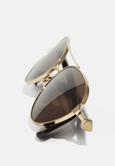 Goldgerahmte Piloten-Sonnenbrille mit dunklen Gläsern, die auf einer weißen Oberfläche liegen, aus einem Winkel betrachtet, der die zusammengefalteten Bügelenden zeigt.