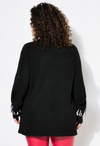 Schwarzer Pullover mit langen Ärmeln und strukturiertem Strick, mit gemusterten weißen Akzenten an den Bündchen. Kombiniert mit rosa eng anliegenden Hosen.