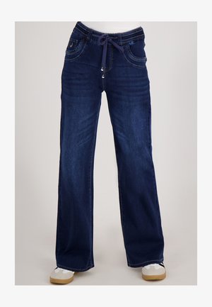 Donkerblauwe wijde jeans met elastische tailleband en marineblauwe trekkoord, gedragen met witte en beige sneakers.