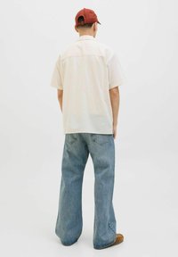 Camisa de botones de manga corta en color crema claro, combinada con jeans de pierna ancha en azul claro y una gorra de béisbol roja, vista desde atrás.