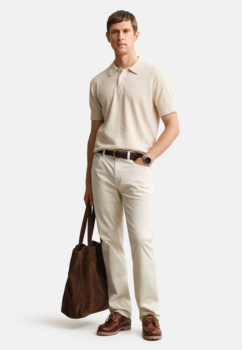 Uomo con polo beige a maniche corte, pantaloni chiari, cintura e scarpe marroni, che tiene una grande borsa tote marrone con una mano in tasca.