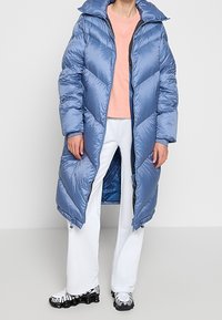 Personne portant un long manteau en duvet bleu, un sweat-shirt pêche, un pantalon blanc et des baskets noir et blanc, se tenant devant un fond uni.