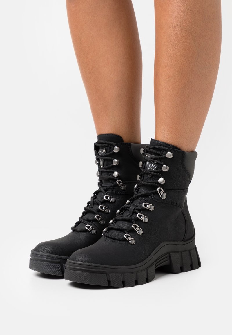 Bottes noires à semelle crantée épaisse, œillets argentés et lacets épais portées sur des jambes nues sur fond blanc.