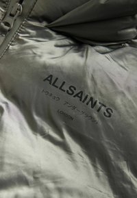 Tessuto imbottito verde oliva con una texture liscia, caratterizzato dal logo stampato nero "ALLSAINTS" e "LONDRA" in inglese e giapponese.
