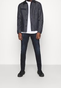 Dark denim jacket with a contrast black collar, front pockets, and black buttons, paired with dark jeans and black sneakers.