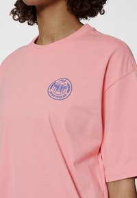 Rosa bomullströja med rund halsringning och korta ärmar. Har en blå cirkulär logotyp med en bi- och lagerkransdesign på bröstet.