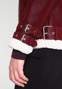 Manche de veste en cuir bourgogne avec doublure en fourrure blanche, dotée de deux boucles ajustables et d'une texture lisse. Vêtements sombres visibles en dessous.