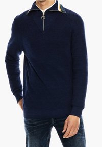 Marinblå stickad tröja med dragkedja i kragen och en randig accent i grönt, gult och rött; strukturerad tyg och avslappnad passform.