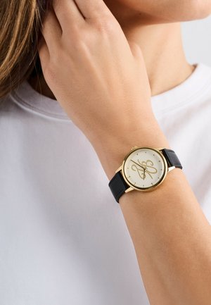 Guldtonad klocka med en rund, vit urtavla som har en logotyp, placerad på ett svart läderarmband. Hand som vilar mot en vit skjorta.