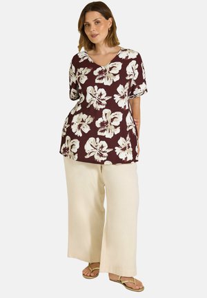 Femme debout, portant un haut bordeaux à motifs floraux, un pantalon crème à jambes larges et des sandales, sur un fond uni clair.