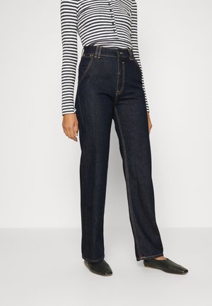 Personne portant un jean large bleu foncé taille haute, des ballerines noires pointues et un haut à manches longues rayé noir et blanc.
