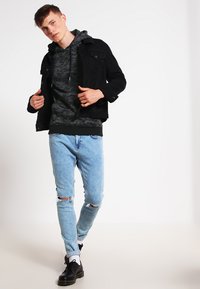 Svart denimjakke over en grå kamuflasje-hettegenser, parret med lyseblå ripped jeans og svarte sko. Nøytral bakgrunn, stående posering.