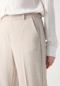 Beige Leinenhosen mit lässiger Passform, seitlichen Taschen und subtiler Textur. Kombiniert mit einer weißen, gerippten Bluse mit langen Ärmeln.