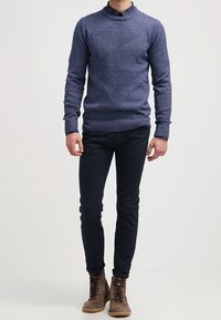 Suéter de punto naval con un patrón de diamante texturizado, puños y dobladillo acanalados, combinado con pantalones oscuros y botas marrones con cordones.