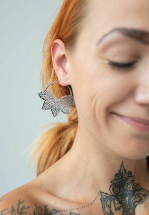 Silver leaf-shaped earrings with intricate cut-out details, hanging from a loop, showcasing a delicate design and fine texture.