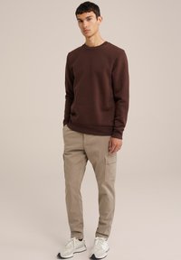 Felpa marrone con scollo rotondo, polsini aderenti e pantaloni cargo beige chiaro. Sneakers bianche e grigie, in piedi su uno sfondo neutro.