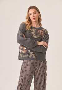 Suéter gris con bordado floral de lentejuelas, escote redondo y hombros caídos, combinado con pantalones de satén con un sutil patrón de hojas.