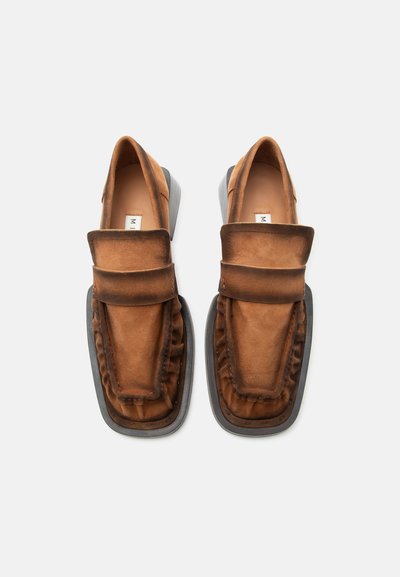 Καφέ σουέτ μοκασίνια με φαρδιά λωρίδα στο πάνω μέρος και μαύρες καουτσούκ σόλες, εμφανιζόμενα από πάνω σε λευκό φόντο.