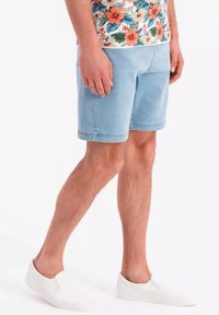 Shorts en denim bleu clair à coupe droite, avec des ourlets légèrement effilochés, portés avec une chemise à motifs floraux et des chaussures blanches sans lacets.