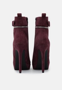 Burgundské vysoké kotníkové boty na jehlovém podpatku z veluru s elegantním designem, vybavené zipem a detaily s přezkou, a kontrastní černou podrážkou.