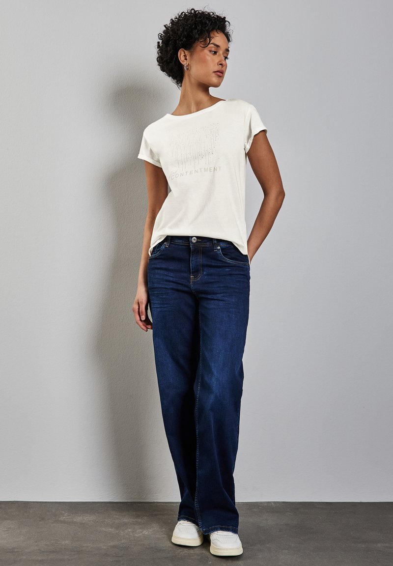 Bílý bavlněný tričko s textovým designem "CONTENTMENT", spárované s tmavě modrými širokými džínami a bílými teniskami. Jednoduchý a moderní outfit.
