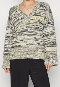 Pull en tricot avec un col en V, coupe oversize et manches longues, dans un motif jaune, noir et blanc. Tissu texturé avec des poignets côtelés.