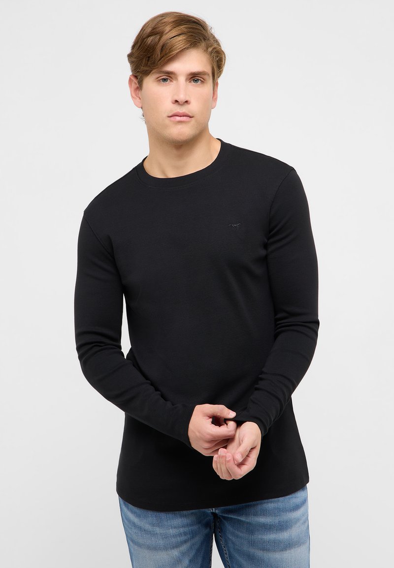 Młody mężczyzna z jasnobrązowymi włosami, ubrany w dopasowaną czarną koszulę z długim rękawem i niebieskie dżinsy, poprawiający rękaw na jednolitym tle.