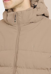 Beige pufferjacka med hög krage, framzipper och justerbar huva. Har en subtil logo och textil med kviltat mönster.