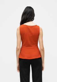 Γυναίκα με μακριά σκούρα μαλλιά, που φοράει πορτοκαλί τοπ χωρίς μανίκια και μαύρο παντελόνι, στέκεται με την πλάτη προς το μέρος μας σε απλό φόντο.