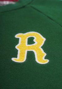 Grön sweatshirt med ett stort, fluffigt gult "R" med vit brodyr som omger bokstaven. Strukturerad tyg med ribbad halsringning.