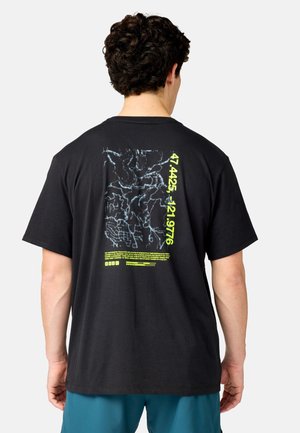 Uomo con capelli corti e ricci veste una t-shirt nera con una grafica astratta bianca e numeri e testi gialli al neon sulla schiena.