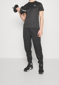 Černé sportovní tričko s vlnkovým vzorem, doplněné o černé tepláky s bočními akcenty a černé sportovní boty, které drží činku.