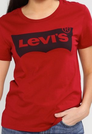 Vrouw draagt een rood Levi's T-shirt met zwart logo, één hand in de spijkerbroekzak, tegen een effen lichte achtergrond.
