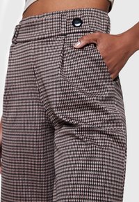 Närbild av en persons hand i fickan på höga, bruna och beige houndstooth-mönstrade byxor med svarta knappar.