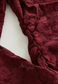 Burgundy spets tyg har ett blommönster med intrikata detaljer och en texturerad yta, draperat och samlat i sektioner.