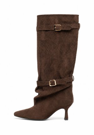 Stivale marrone in suede fino al ginocchio con punta affusolata, due cinturini decorativi con fibbie dorate e tacco sottile di altezza media.