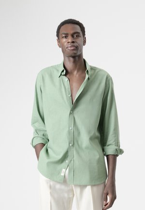 Hombre con cabello corto y rizado que lleva una camisa verde claro abotonada con las mangas remangadas y pantalones blancos, de pie frente a un fondo liso.