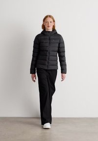 Black puffer jacket with horizontal stitching, a hood, and a zip front closure. Worn over black pants and white sneakers, standing on a neutral floor.