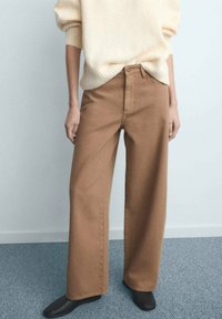 Pantalon large marron en tissu robuste avec une taille haute, associé à un pull crème, sur un fond clair.