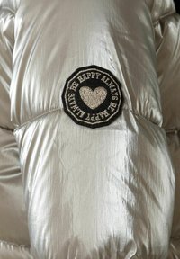 Doudoune argentée en tissu brillant et texturé. Patch noir notable avec un cœur brodé et le texte "SOIS TOUJOURS HEUREUX."