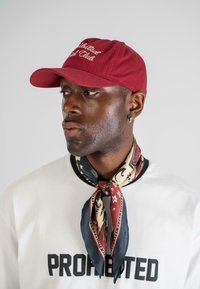 Căciulă de baseball bordeaux cu inscripție albă brodată, asociată cu un eșarfă din mătase cu un model în nuanțe de burgundy, negru și crem, purtată peste o cămașă albă.