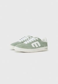 Baskets en suède vert avec des accents blancs, présentant un logo central, une pointe arrondie et des lacets blancs sur une semelle texturée.