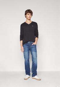 Joven vestido con un suéter negro de cuello en V, jeans azules y zapatillas blancas, de pie con una mano en el bolsillo frente a un fondo liso.