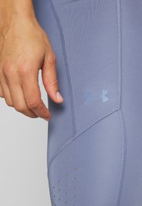 Svetlo modre športne legice z gladko teksturo, ki vključujejo izrazit logotip Under Armour, podrobnosti šivanja in majhne perforacije blizu roba.