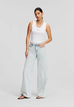 Weißes, geripptes Tanktop mit kleiner blauer Schrift, kombiniert mit hellen, weit geschnittenen Jeans und flachen Sandalen. Das Outfit zeichnet sich durch eine lockere Passform aus.