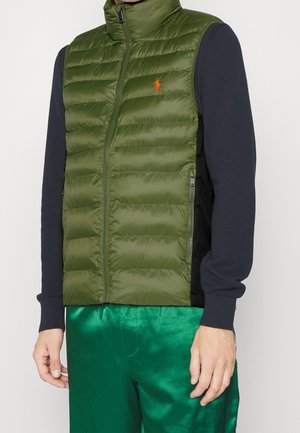Homme portant une doudoune sans manches vert olive avec un logo orange sur un haut à manches longues bleu marine et un pantalon vert brillant sur fond blanc.